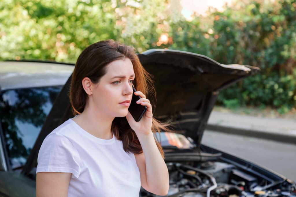 Woman calling for roadside assistance while standing next to broken-down car with open hood.