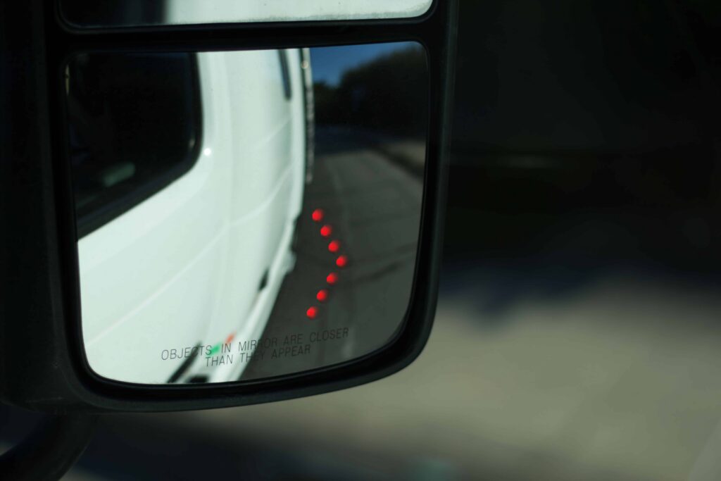Close-up of a truck side mirror with blind spot warning lights, highlighting commercial truck safety and driver visibility on the road.