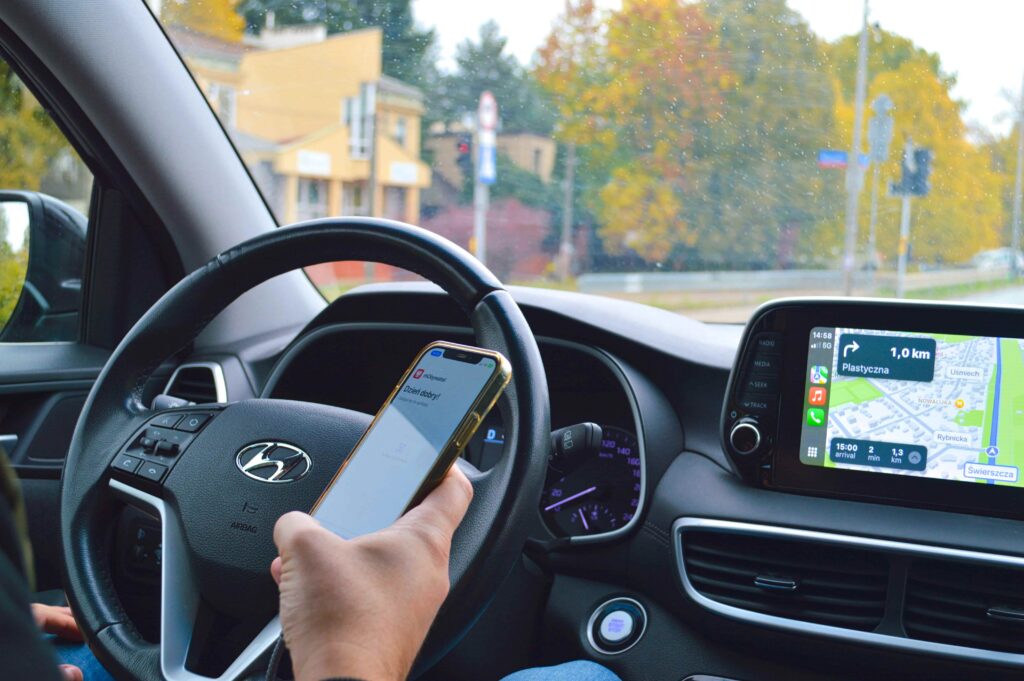 Driver using a smartphone while driving a car with GPS navigation on dashboard, illustrating distracted driving and road safety risk.