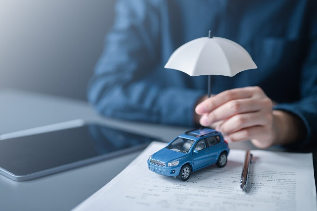 A person holds a small white umbrella over a blue toy car sitting on insurance documents, symbolizing the legal protection offered by San Antonio Uninsured Motorist Accident Lawyers.