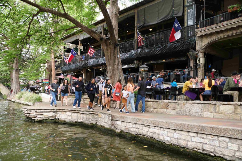 People walk along the San Antonio River Walk past outdoor restaurant patios with Texas flags, representing the local area served by San Antonio Uninsured Motorist Accident Lawyers.