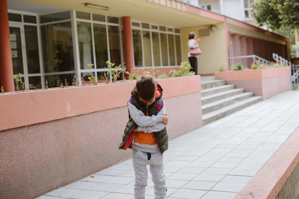 A young boy stands outside a school building with his arms crossed and head down, highlighting options for suing a Texas daycare for child injury negligence.