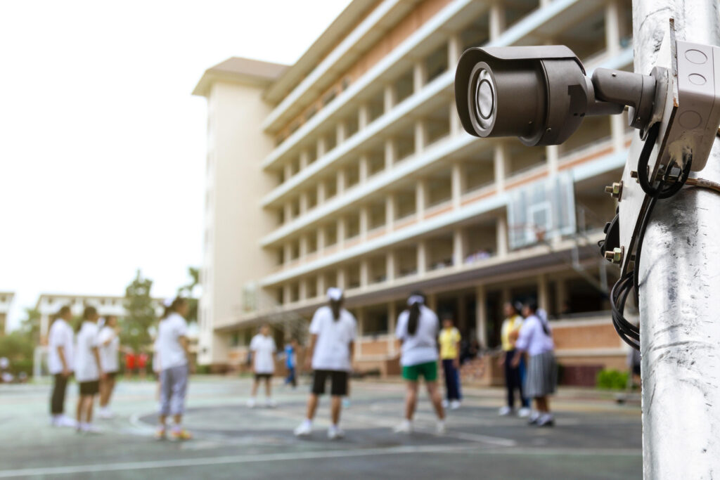 A security camera mounted on a pole overlooks a school courtyard where children play, referencing evidence collection when suing a Texas daycare for child injury negligence.