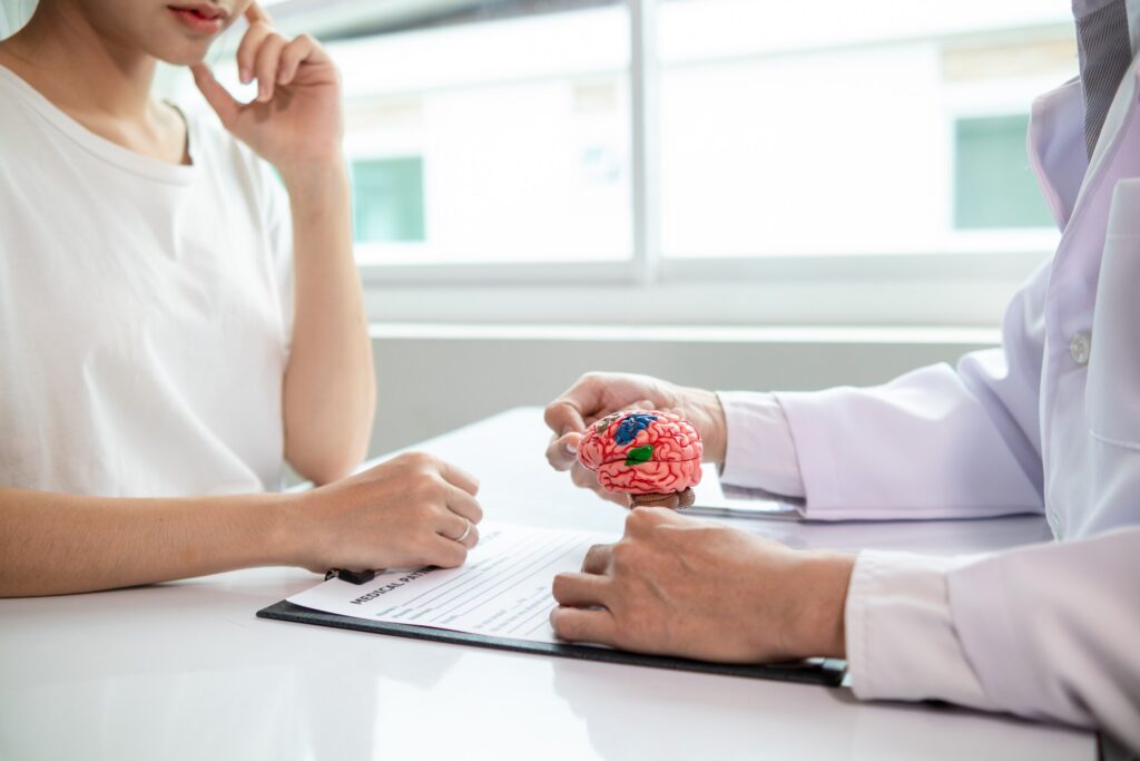 Close-up of a small human brain model on a desk between a doctor and a patient, discussing the lifetime cost of a TBI in Texas & your claim.
