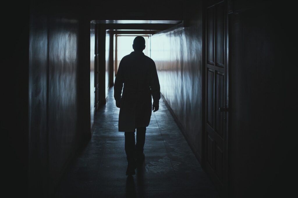 Silhouetted doctor in a lab coat walking down a dimly lit hospital hallway.