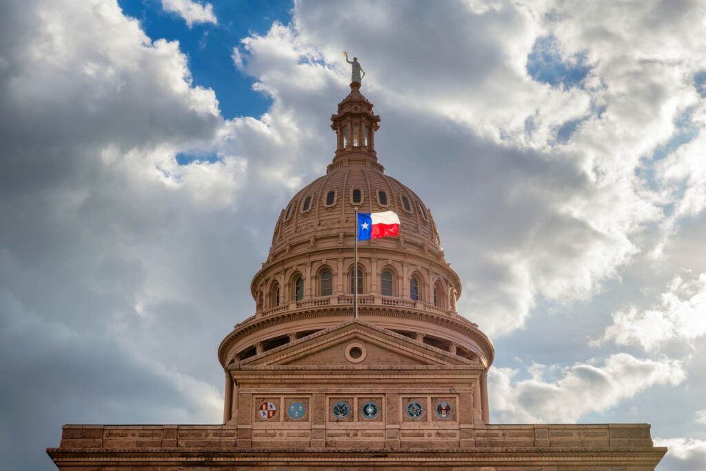 Dome of the Texas State Capitol in Austin with the Texas state flag flying under cloudy skies.